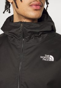 Svart huva jacka gjord av lättviktsmaterial, har en hel dragkedja och en vit logotyp på vänster bröstområde. Texturerad look på tyget.