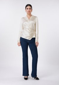 Κρεμ σατέν πουκάμισο τύπου wrap με μακριά μανίκια και λεπτομέρεια με δέσιμο, συνδυασμένο με σκούρα μπλε τζιν καμπάνα και μαύρες γόβες με μυτερή μύτη.