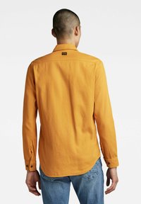 Camicia a maniche lunghe di un brillante arancione, realizzata in tessuto morbido. Presenta un colletto e polsini con chiusure a bottone. Sul retro è visibile un'etichetta con il logo.