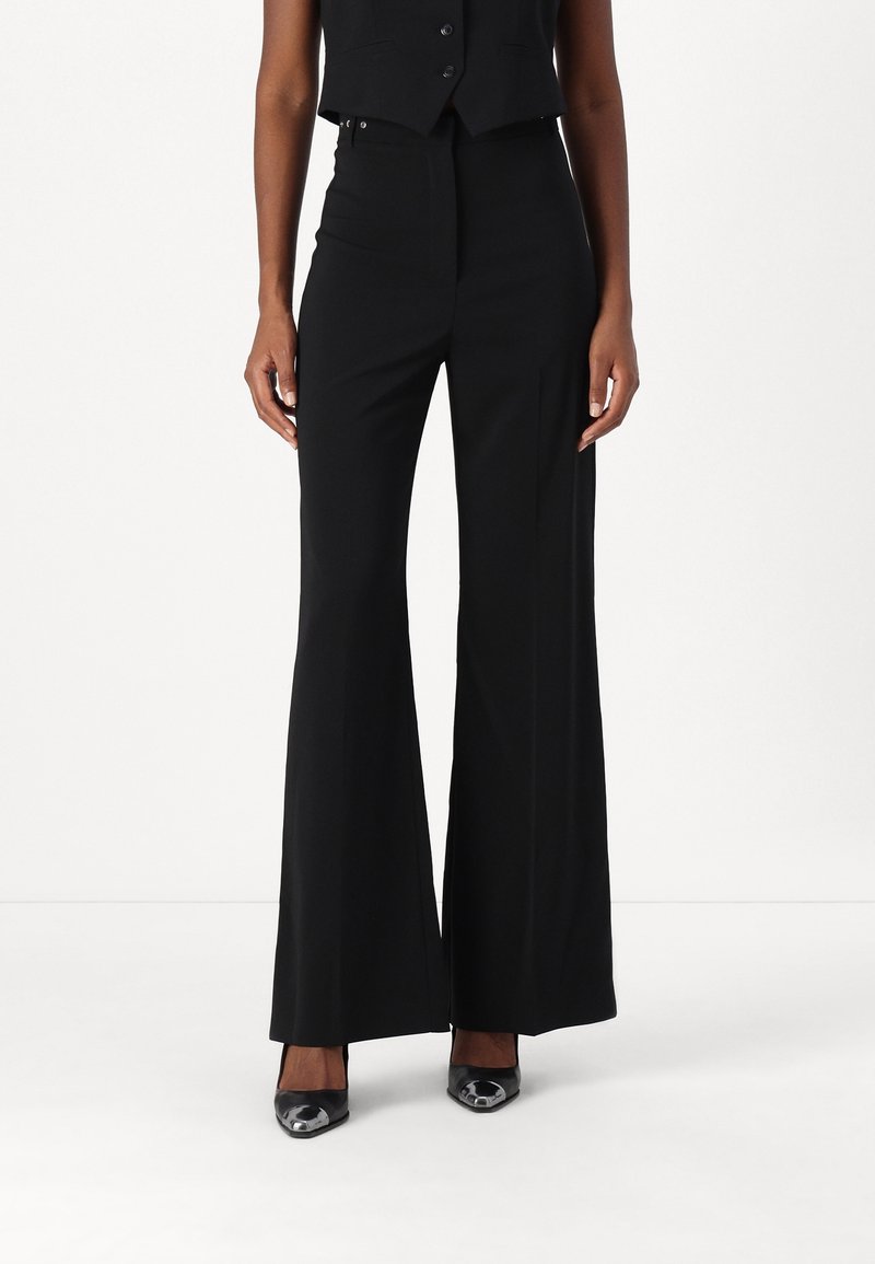 Femme portant un pantalon noir taille haute à jambes larges, un gilet noir sans manches boutonné, et des talons noirs pointus, debout devant un fond blanc.