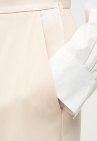 Χέρι γλιστράει στην τσέπη μπεζ παντελονιού, φορώντας λευκό πουκάμισο με μακριά μανίκια και ορατό μανσέτα.