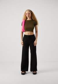 Top corto verde, pantalones de pierna ancha negros y sandalias negras gruesas. Accesorio rosa de peluche colgado sobre un hombro. Gafas de sol con marco transparente.