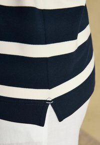 Marineblaue und weiße gestreifte Saumlinie eines Hemdes mit einem kleinen seitlichen Schlitz, getragen über weißen Hosen vor neutralem Hintergrund.