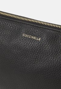 Pochette en cuir noir avec une surface texturée, une fermeture éclair dorée et un logo doré portant le nom "COCCINELLE" à l'extérieur.