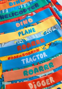 Sous-vêtements pour enfants colorés empilés, chaque ceinture porte une étiquette avec des mots comme "HÉLICOPTÈRE", "DINO", "AVION", "ROARR", "TRACTEUR" et "PELLETEUSE".