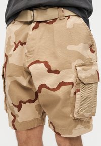 Μπεζ και καφέ cargo σορτς παραλλαγής με πλαϊνή τσέπη και υφαντή ζώνη, φορεμένα με μαύρο πουκάμισο, μερική θέα σε πόδια και μέση.