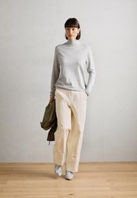 Svijetlosivi dukserica s dugim rukavima, bež široke hlače i obuća s uzorkom; model drži maslinasto zelenu jaknu. Minimalistička pozadina.