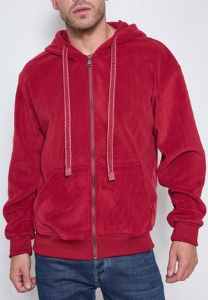 Homme portant un sweat à capuche rouge zippé avec cordons de serrage et poches avant, associé à un jean bleu, sur un fond uni.