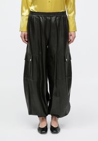 Άτομο που φοράει μαύρο δερμάτινο παντελόνι φαρδιάς γραμμής με τσέπες cargo, συνδυασμένο με μαύρα μπαλαρίνες και κίτρινο σατέν πουκάμισο με μακριά μανίκια.