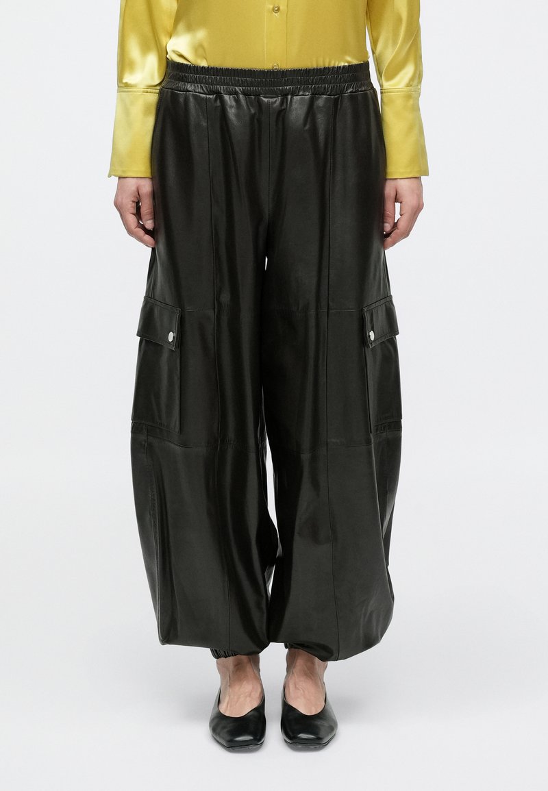 Άτομο που φοράει μαύρο δερμάτινο παντελόνι φαρδιάς γραμμής με τσέπες cargo, συνδυασμένο με μαύρα μπαλαρίνες και κίτρινο σατέν πουκάμισο με μακριά μανίκια.