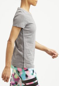 Žena ve středním kroku v šedém sportovním tričku s krátkým rukávem a barevných legínách s černými vodorovnými pruhy.
