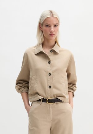 Blondynka w beżowej kurtce zapinanej na guziki z podwiniętymi rękawami oraz dopasowanych, wysokich spodniach z rękami w kieszeniach, neutralne tło.