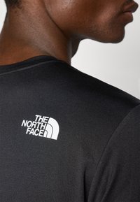 T-shirt de performance noir avec une texture lisse, présentant un logo blanc "The North Face" à l'arrière près de l'encolure.