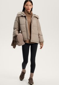 Giacca a vento con imbottitura leggera, design trapuntato, con cappuccio rimovibile. Il modello indossa un maglione marrone, leggings neri e sneakers marroni.