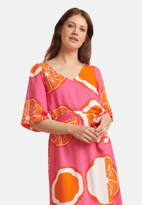Rosa Kleid mit einem auffälligen orange-weißen Fruchtmuster, mit V-Ausschnitt, kurzen Puffärmeln und einem Taillenzug. Glatter Stoff.