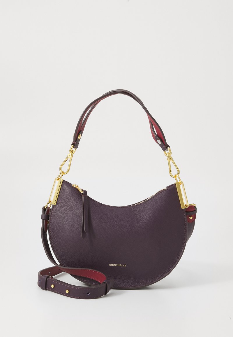 Purple leather handbag with a curved shape, gold hardware, detachable strap, and textured surface. Logo displayed on the front.