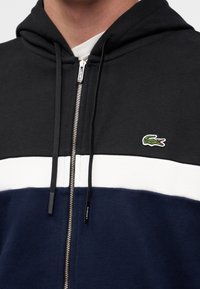 Homme portant un sweat à capuche zippé color-block noir, blanc et marine avec un logo crocodile vert sur la poitrine et des cordons sur la capuche.