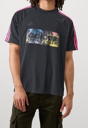 Man draagt een zwart Adidas sportshirt met roze strepen op de schouders en een meerkleurig Adidas-logo op de borst, gecombineerd met donkergroene broek.