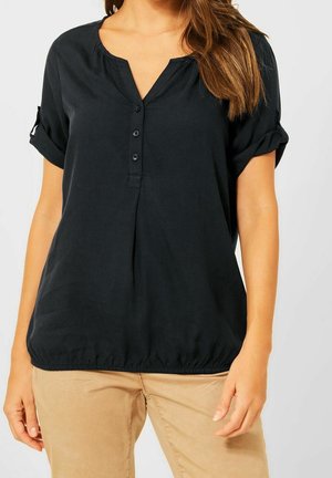 Femme portant un chemisier noir à manches courtes boutonné avec des poignets retroussés et un pantalon beige, debout devant un fond uni.