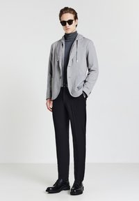 Giovane uomo che indossa occhiali da sole neri, felpa grigia sotto un blazer grigio, pantaloni neri e scarpe eleganti nere su uno sfondo bianco uniforme.