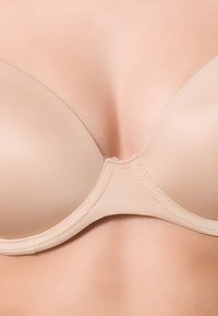 Beige gewatteerde bh met een gladde stofafwerking, voorzien van een diepe halslijn en subtiele naden voor ondersteuning, ontworpen voor een slanke laag onder kleding.