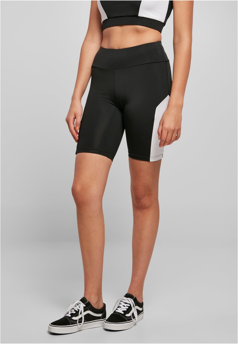 Pantalones cortos deportivos en blanco y negro hechos de material elástico, con una cintura alta y panel lateral de colores contrastantes. Usados con zapatillas negras.