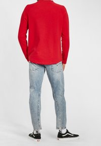 Chemise rouge à manches longues, jean bleu clair déchiré, et baskets noires avec semelles blanches. Le tissu semble doux ; le jean possède des poches arrière.