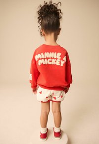 Punainen huppari, jossa lukee "MINNIE & MICKEY" pörröisillä valkoisilla kirjaimilla, yhdistettynä valkoisiin shortseihin, joissa on punaisia sydänkuvioita, ja punaisiin kenkiin.