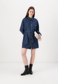 Abito camicia in denim blu scuro, con bottoni sul davanti, colletto e vita con fiocco. Tasche sul petto e maniche corte. Stivali neri alla caviglia.