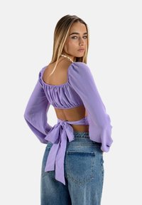 Lilla cropped bluse med pufærmer og en bindebag detalje, parret med højtaljede denim jeans. Glat stof med en let tekstur.