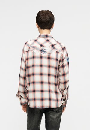 Asmuo dėvi rožinę, baltą ir tamsiai mėlyną languotą ilgomis rankovėmis marškinius, su sparnuoto, apskritos formos logotipo pleistru viršutinėje nugaros dalyje, atsukęs nugarą, baltame fone.