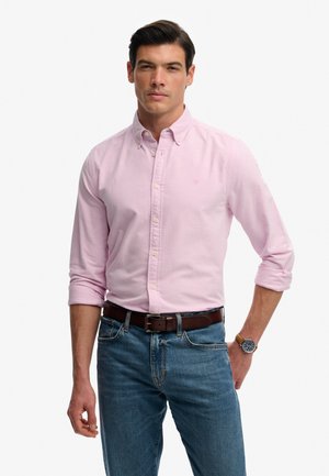 Uomo che indossa una camicia rosa chiaro con bottoni, maniche arrotolate, jeans blu, cintura marrone e un orologio con cinturino in pelle marrone.