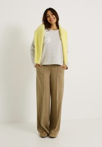Sudadera gris con bordado floral blanco, combinada con pantalones de pierna ancha en beige. Un suave chal amarillo cae sobre el hombro. Calzado marrón.