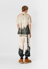 Camiseta gráfica beige y negra y pantalones a juego con estampado de paisaje; los pantalones presentan un logo y texto vertical; modelo lleva botas marrones.