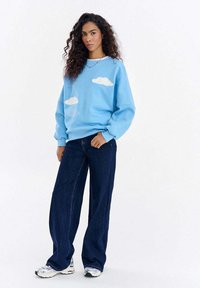 Jasnoniebieska bluza z wzorami chmur, dzianinową fakturą i haftowanym napisem. Do tego szerokie spodnie z ciemnego denimu i białe trampki.