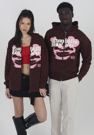Brune zip-up hettegensere med rosa grafisk tekst, båret av to modeller. Den ene har på seg et svart skjørt og glansfulle støvler til knærne, mens den andre har lyse bukser.
