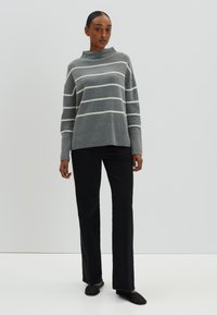 Pull gris rayé avec un col montant et un ourlet côtelé, associé à un pantalon noir à jambe droite et des chaussures plates dans un cadre sobre.