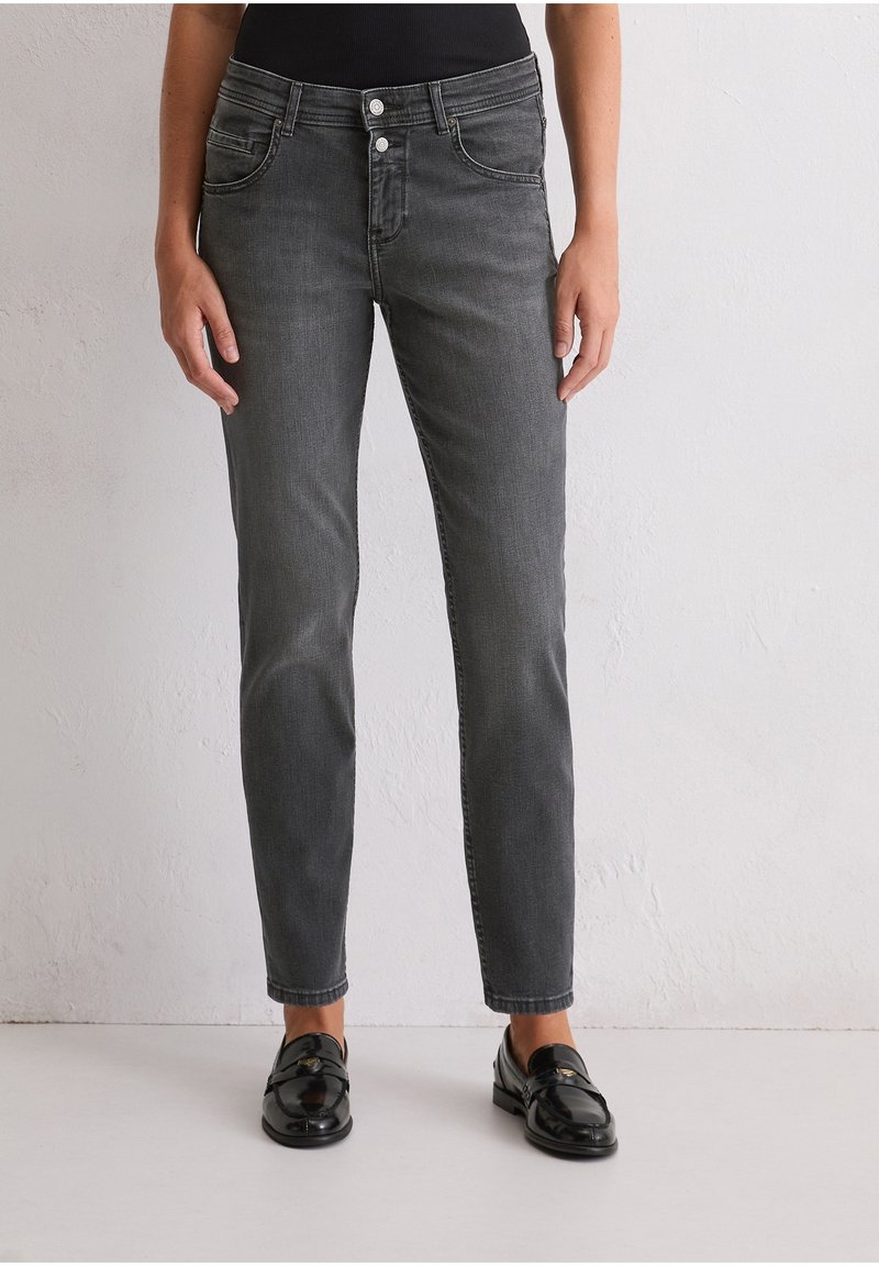 Personne portant un jean slim gris foncé et des mocassins noirs brillants, debout devant un mur clair uni.