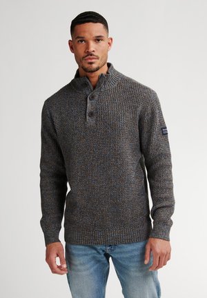 Hombre con un suéter de punto texturizado en tonos marrón y azul, con cuello abotonado y jeans de color azul claro, de pie contra un fondo liso.