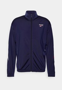 Tmavě modrá bunda Reebok z hladkého, lehkého materiálu. Má vysoký límec, celopropínací zip a kontrastní bílé a červené detaily.