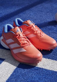 Bright orange athletic shoes with blue accents, featuring a mesh upper, three white stripes, textured sole, and pointed traction studs.