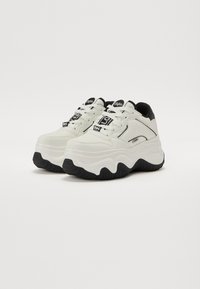 Weiße klobige Sneakers mit einer strukturierten Gummisohle, schwarzen Akzenten und Schnürdesign. Mit Logo auf der Zunge und gemusterten Details an den Seiten.
