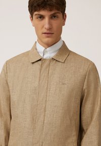 Chaqueta de mezcla de lino beige con cuello con solapa, tapeta delantera y un sutil detalle del logo de perro. Textura suave, ligera y con un ajuste entallado.