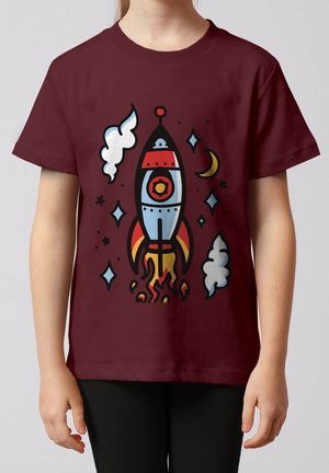 Kind trägt ein bordeauxrotes T-Shirt mit einer bunten Rakete, die zwischen Sternen, Wolken und einem Halbmond startet.
