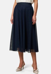 Jupe en tulle marine avec une taille ajustée, un design évasé et une superposition transparente, associée à des escarpins beige à talons ouverts et un accent en nœud.