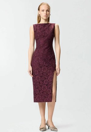 Mujer con vestido midi de encaje burdeos sin mangas con abertura lateral y zapatos de tacón bajo con estampado, de pie contra un fondo claro.