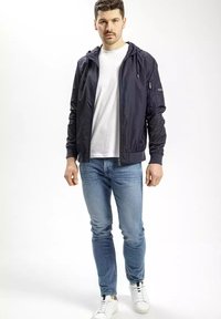 Granatowa kurtka z kapturem, zapinana na zamek, z elastycznymi mankietami i kieszeniami bocznymi, noszona na białym T-shircie i niebieskich dżinsach, w połączeniu z białymi sneakersami.