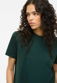 Maglietta in cotone verde scuro con maniche corte, colletto tondo e texture morbida, caratterizzata da una vestibilità rilassata e senza motivi o dettagli visibili.