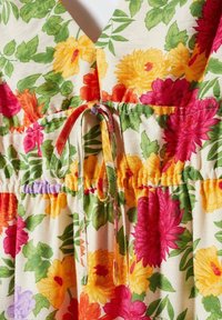 Robe florale avec un décolleté en V, une taille froncée et un détail de nouage. Fleurs colorées en rose, rouge, jaune et vert sur un fond crème.