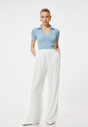 Mujer de pie con las manos en los bolsillos, vistiendo un top de punto azul claro de manga corta y pantalones anchos blancos de cintura alta sobre un fondo liso.